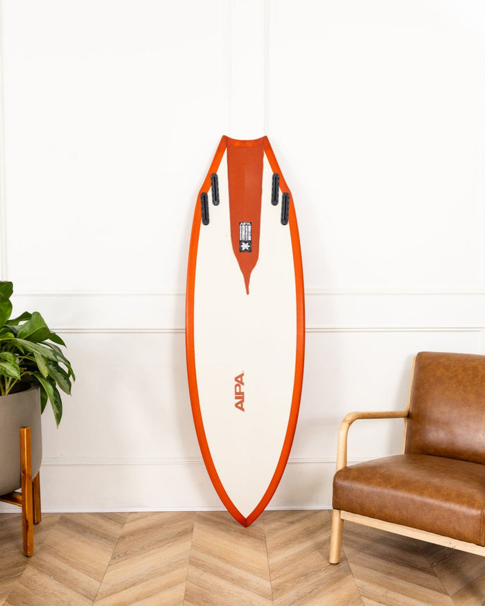 Orange and white surfboard leaning against a white wall with a brown leather chair and potted plants in the foreground.